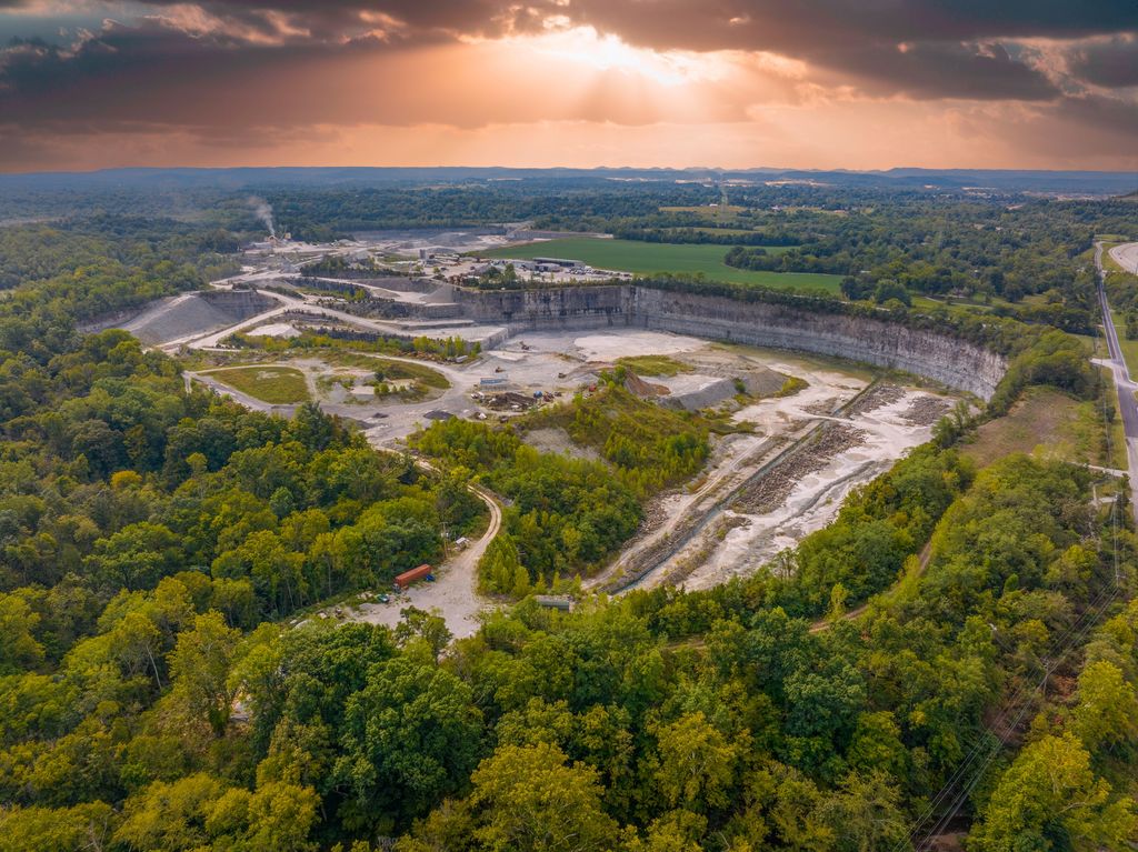 Sun breaking through clouds over a large quarry pit surrounded by forest